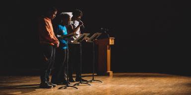 三个黑人学生, 穿便服, 站在图像的右侧, 面向图像中心的,在黑人音乐的背后是一个温暖的木头舞台. 它们被聚光灯照亮, 除了他们自己，几乎所有人都是这样, 舞台的一部分, 还有阴影中的木制讲台. 